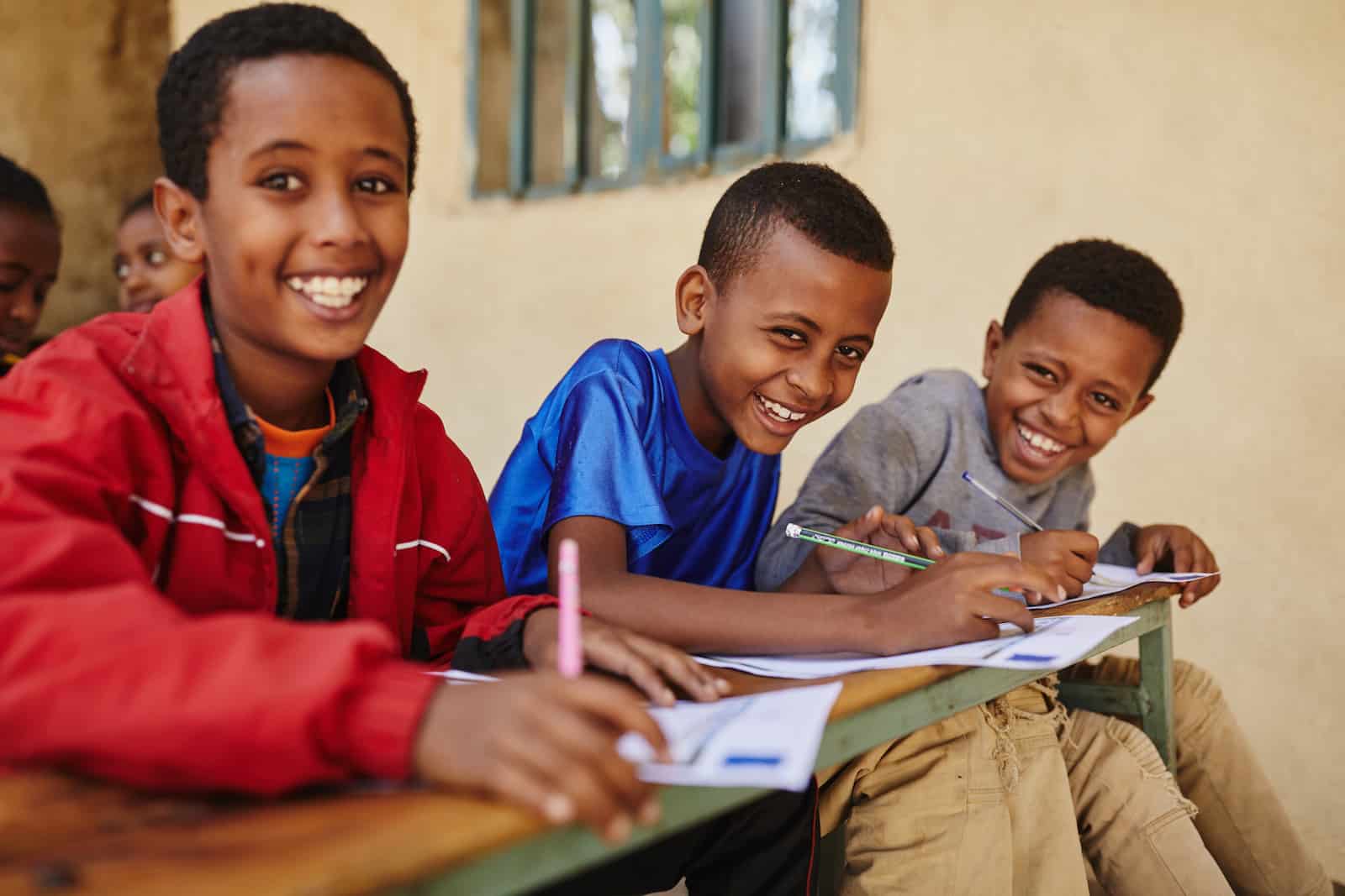 Three boys sit in a row at a desk outside, each writing a letter. They all smile at the camera.