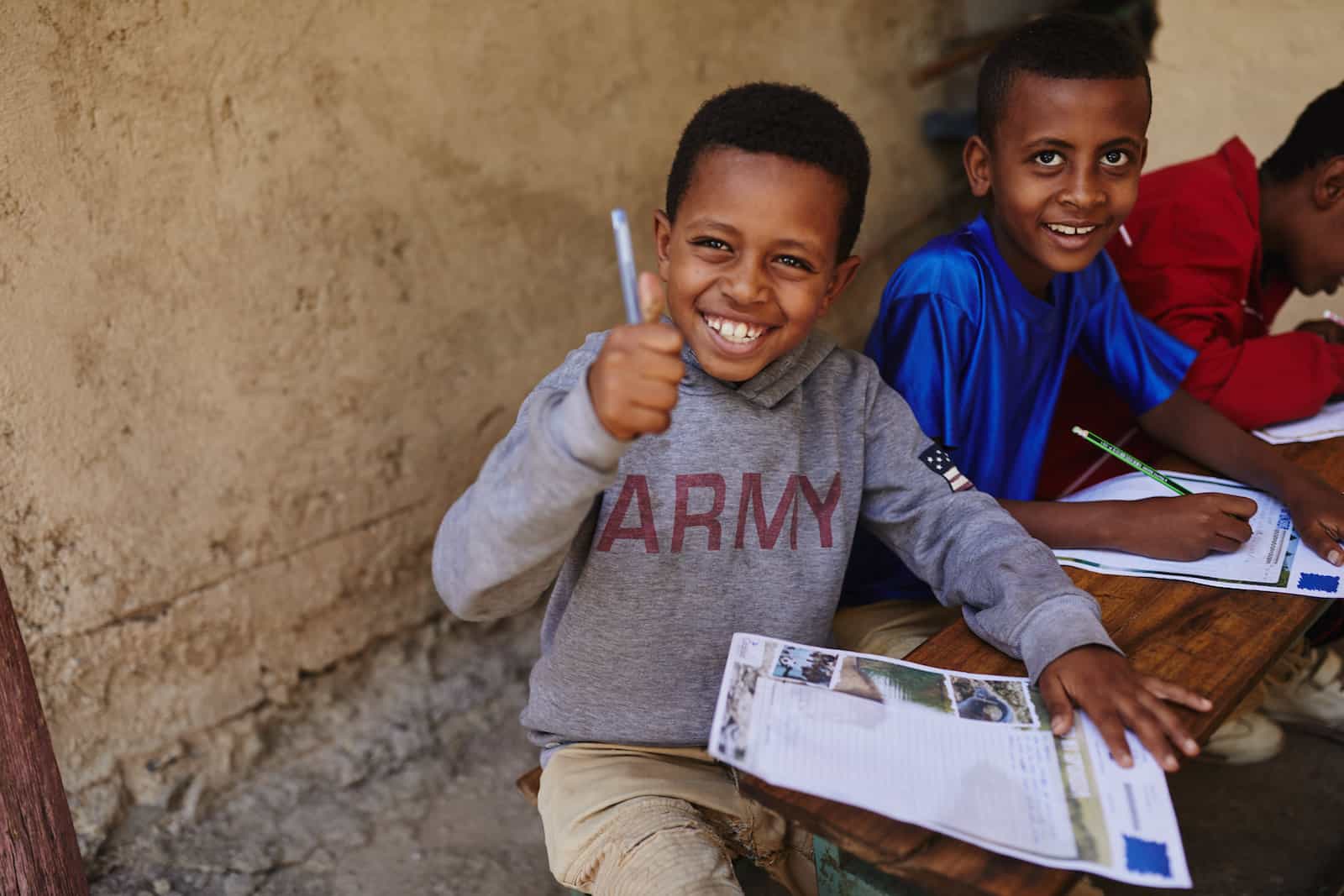 A boy wearing a grey sweatshirt that says "Army" gives the thumbs up sign while holding a pen and paper.