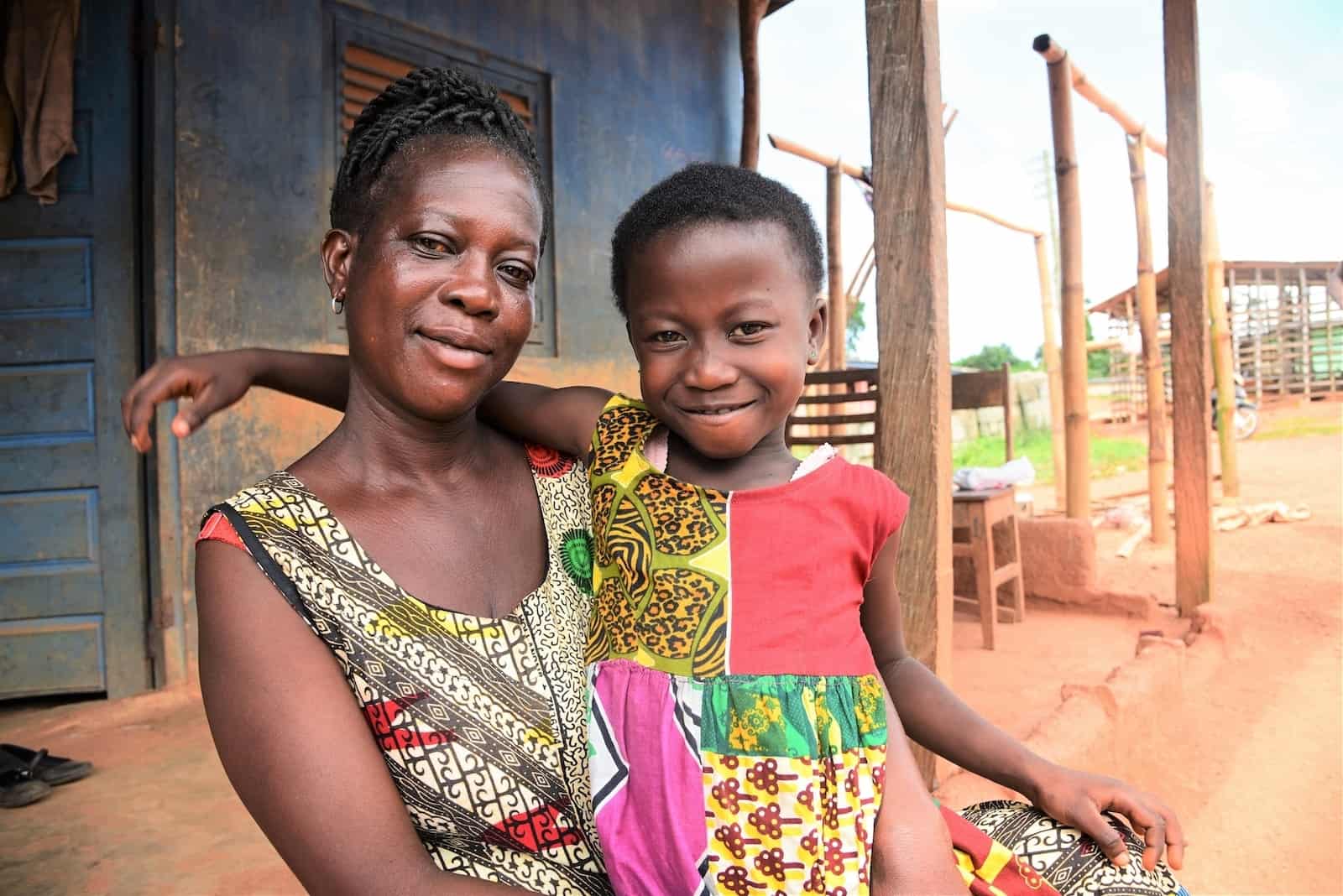 A woman who kept her daughter inside due to a Ghana superstition wears a green and red dress, holding a young girl in her arms in front of a blue house. 