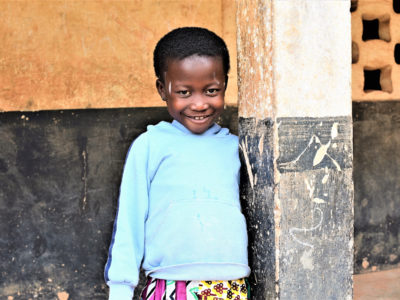 A girl who was trapped inside by a Ghana superstition wears a light blue sweatshirt and patterned dress smiles, standing next to a column in front of a black and white wall.