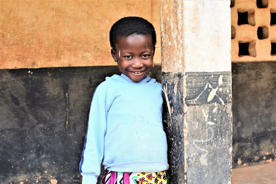 A girl who was trapped inside by a Ghana superstition wears a light blue sweatshirt and patterned dress smiles, standing next to a column in front of a black and white wall.