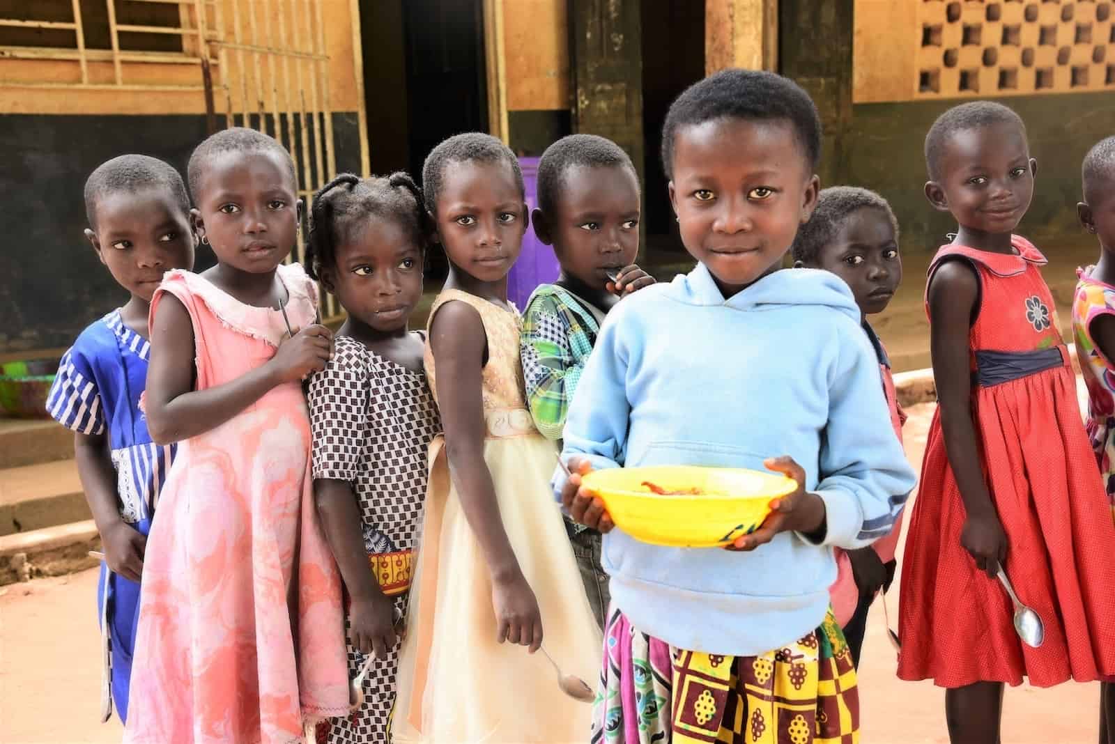 A young girl in a blue sweatshirt and patterned skirt holds a yellow bowl of food. A line of girls stands behind her in line, outside. 