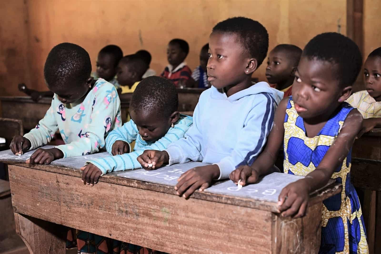 A group of young children sit at a long wooden desk, writing on small chalkboards, looking up at the front of the class. 