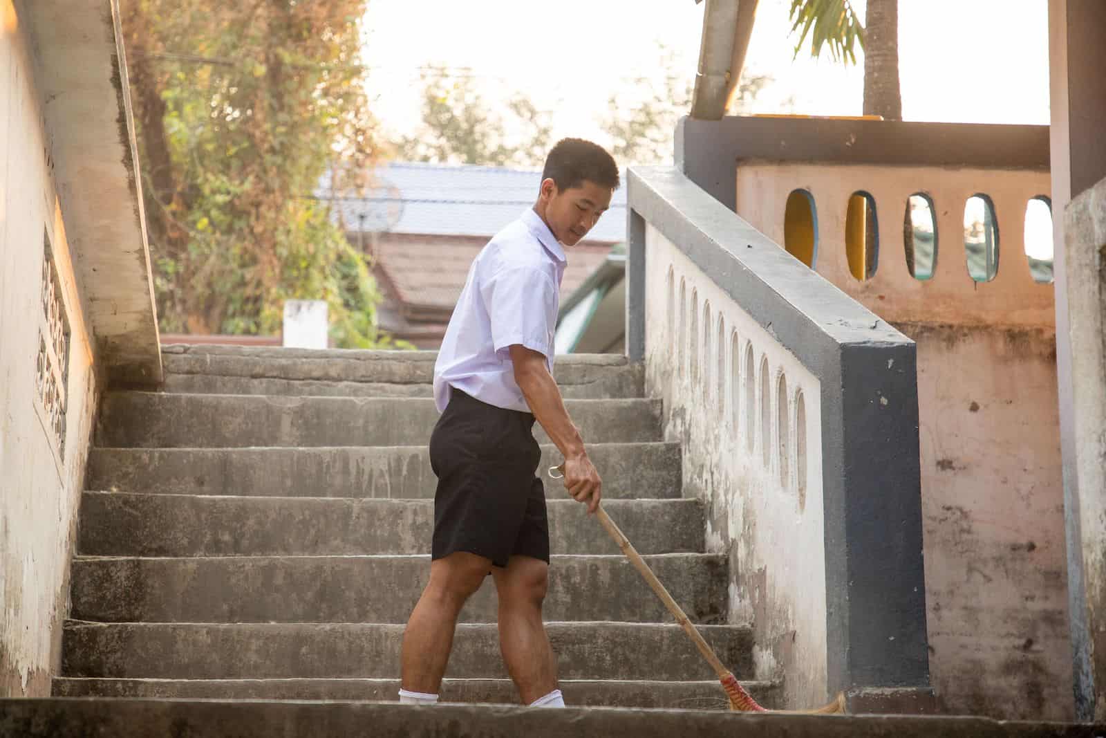 A boy sweeps the stairs outside at a concrete building with trees in the background.
