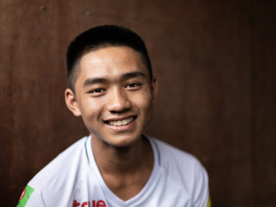 A teenage boy wearing a white shirt smiles at the camera. He sits in front of a brown background.