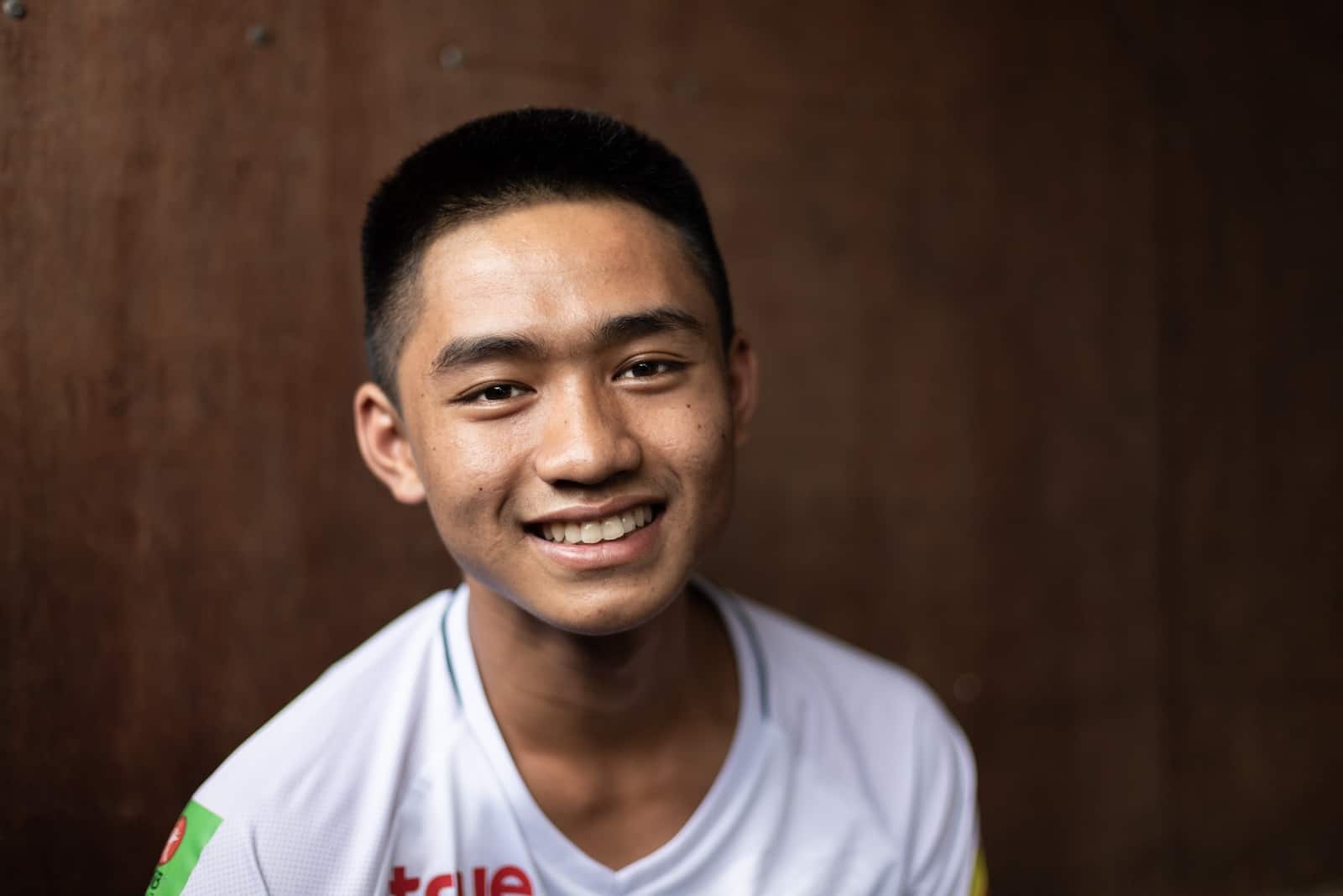 A teenage boy, Adun who was part of the Thailand cave rescue, wears a white shirt smiles at the camera. He sits in front of a brown background. 