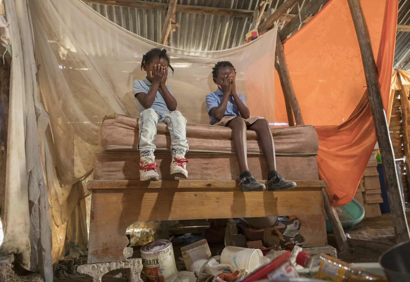 Two girls sit on a bed with their faces covered in prayer. The bed is raised up with concrete blocks. A mosquito net hangs above the bed and the roof is made from corrugated metal. 