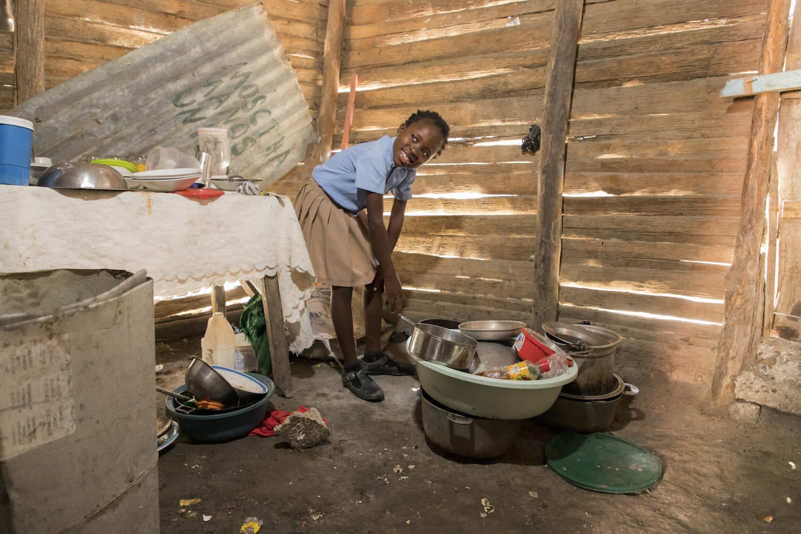 A girl in a blue shirt and tan skirt leans over pots and pans on a dirt floor. There is a table to one side. The walls are made of wood slats with holes in them. 