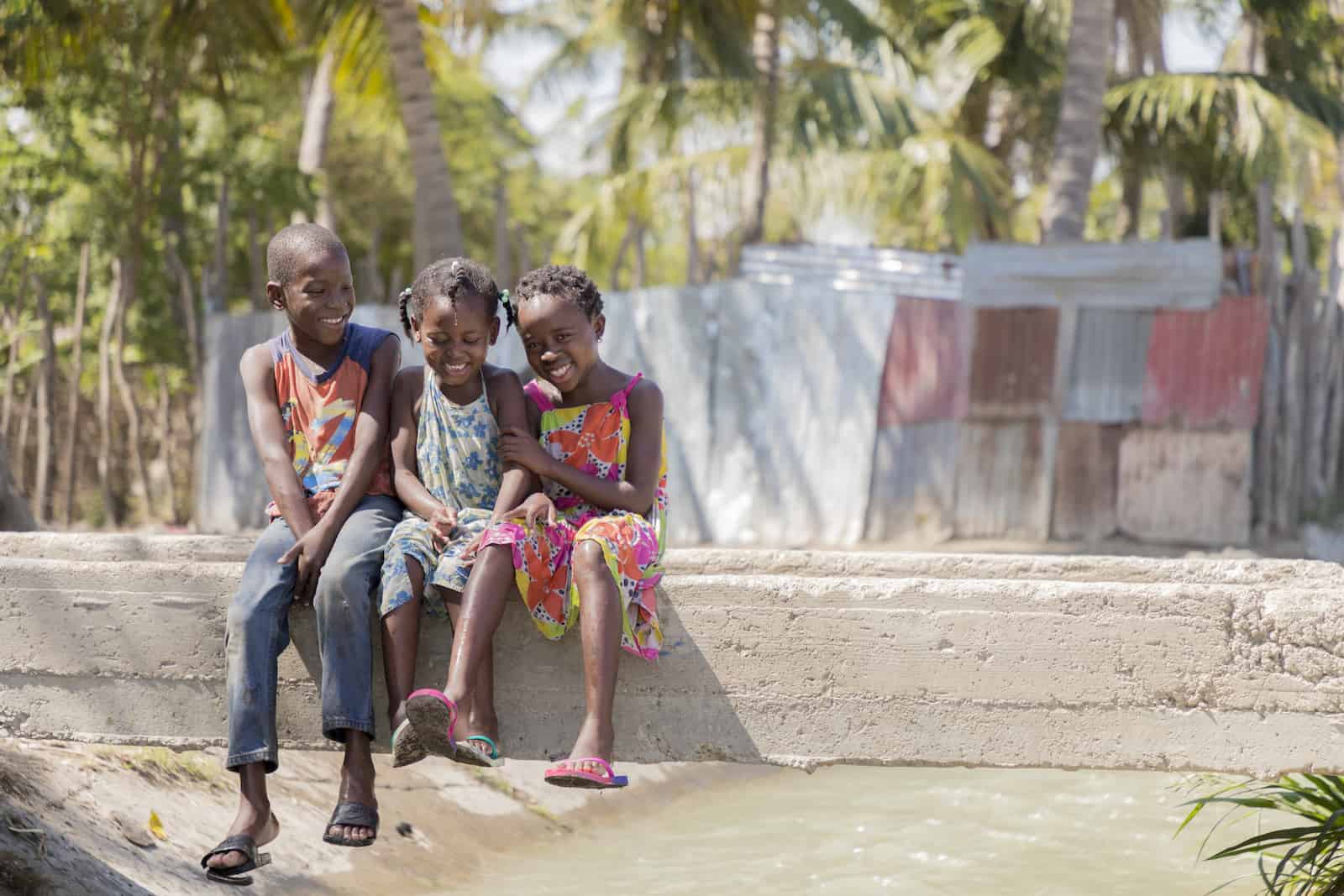 Three young children sit on a concrete beam that is a bridge across a ditch full of water. In the background are palm trees and corrugated metal sheet homes.