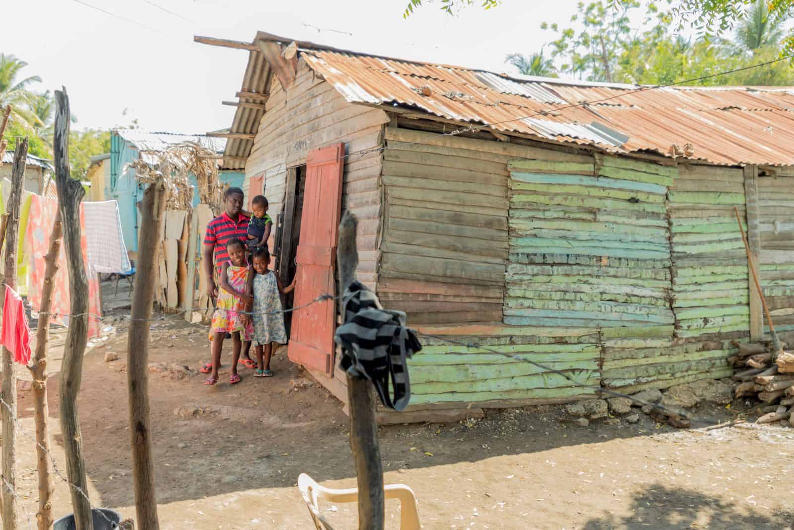 A man stands with three children outside a worn wooden home painted light green. There is a wooden fence in the foreground. 