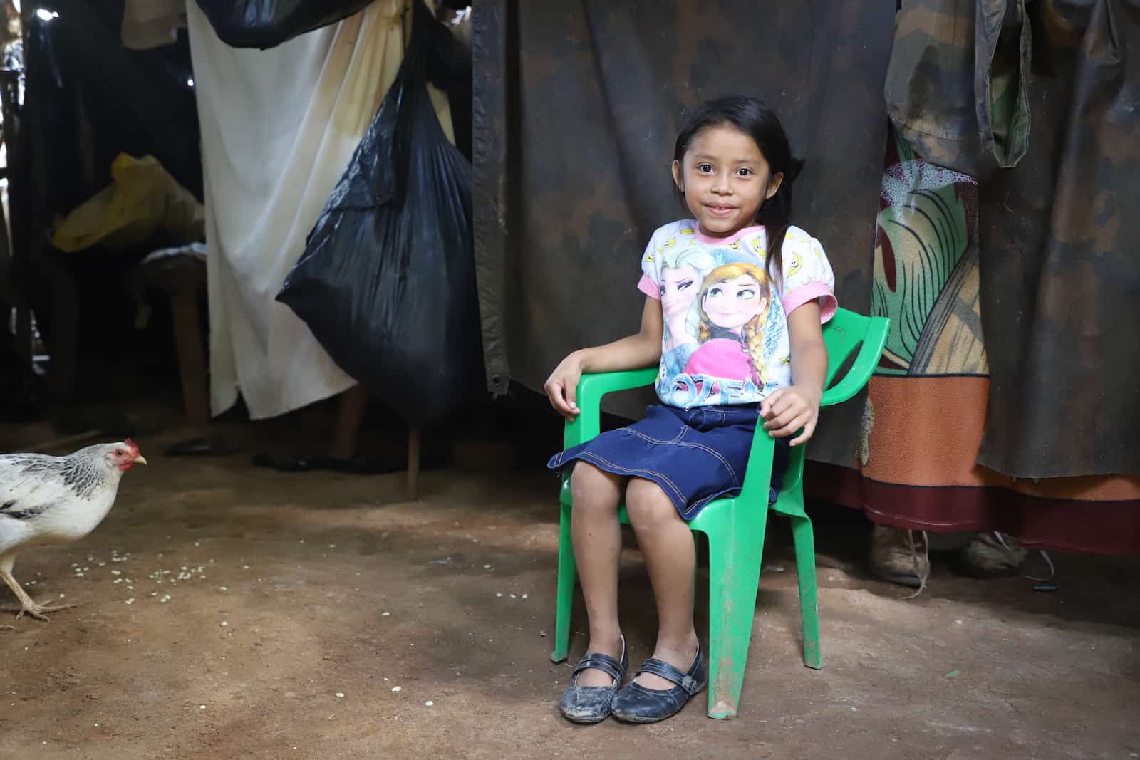 A girl in an "Anna and Elsa" shirt and jean skirt sits in a green plastic chair inside her home in Central America. There are large plastic tarps hanging from the ceiling to serve as walls. A chicken eats corn on the floor. 