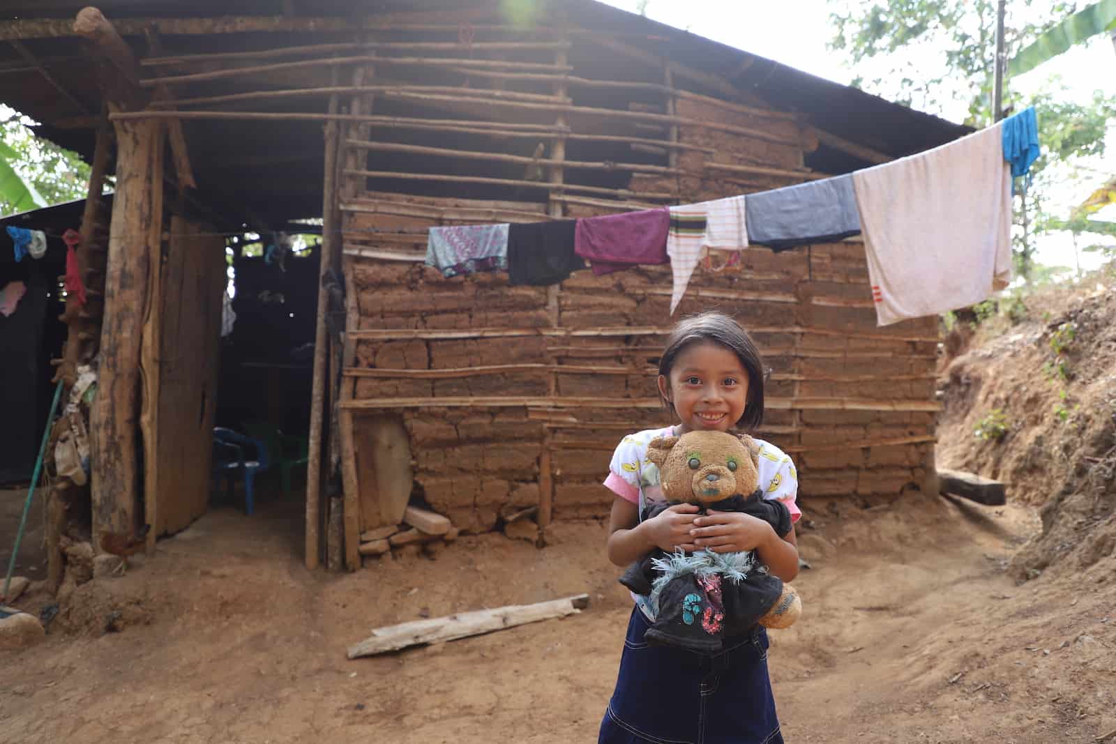 A girl holds a teddy bear, standing in front of a home in Central America made with mud and sticks. There are many holes in the walls. A laundry line hangs out front. 
