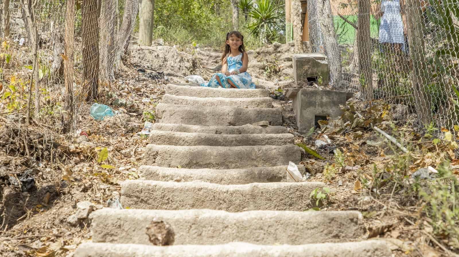 A girl in a blue and white dress sits at the top of steep concrete stairs with chain metal fence on either side. 