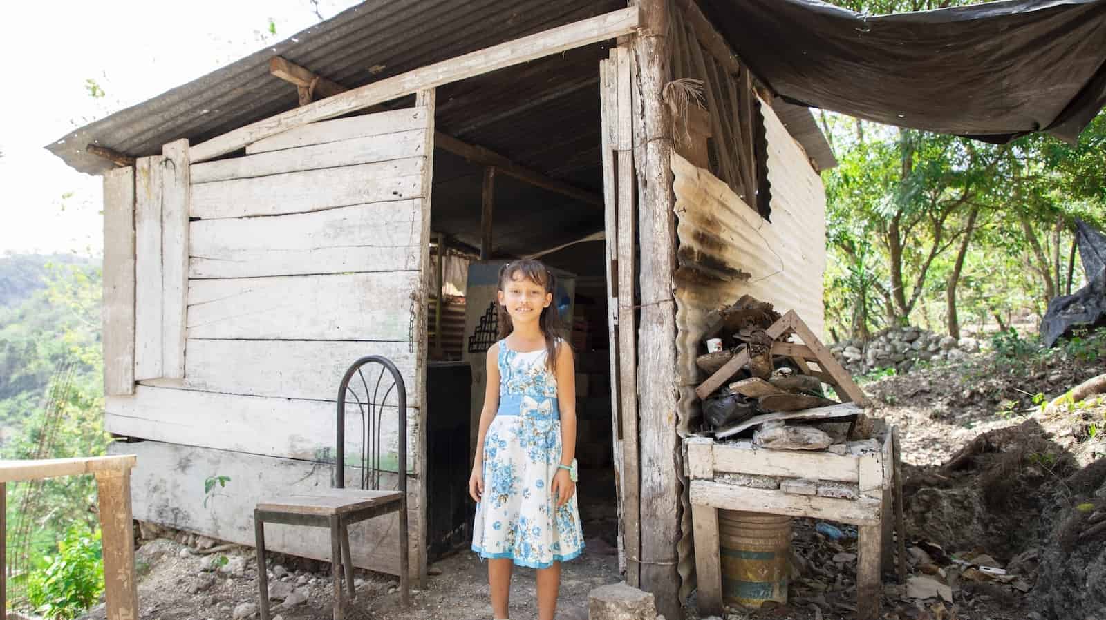A girl in a white and blue dress stands in front of a small home in Central America made from wood and metal sheets. In the background are green hills and tress.