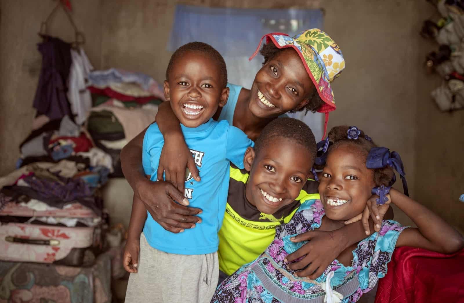 A woman wearing a hat smiles largely, with her arms around three smiling young children. They are inside a concrete home with a stack of clothing in the corner.