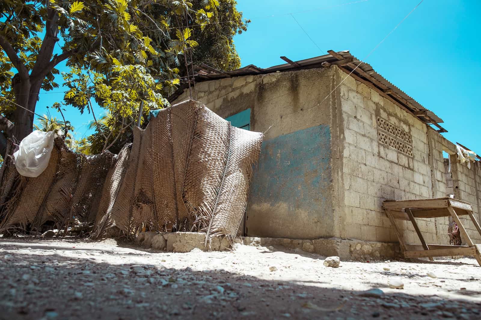 A concrete brick home with a corrugated metal roof stands on a dirt lot surrounded by a woven fence. There is a large tree behind the home. 