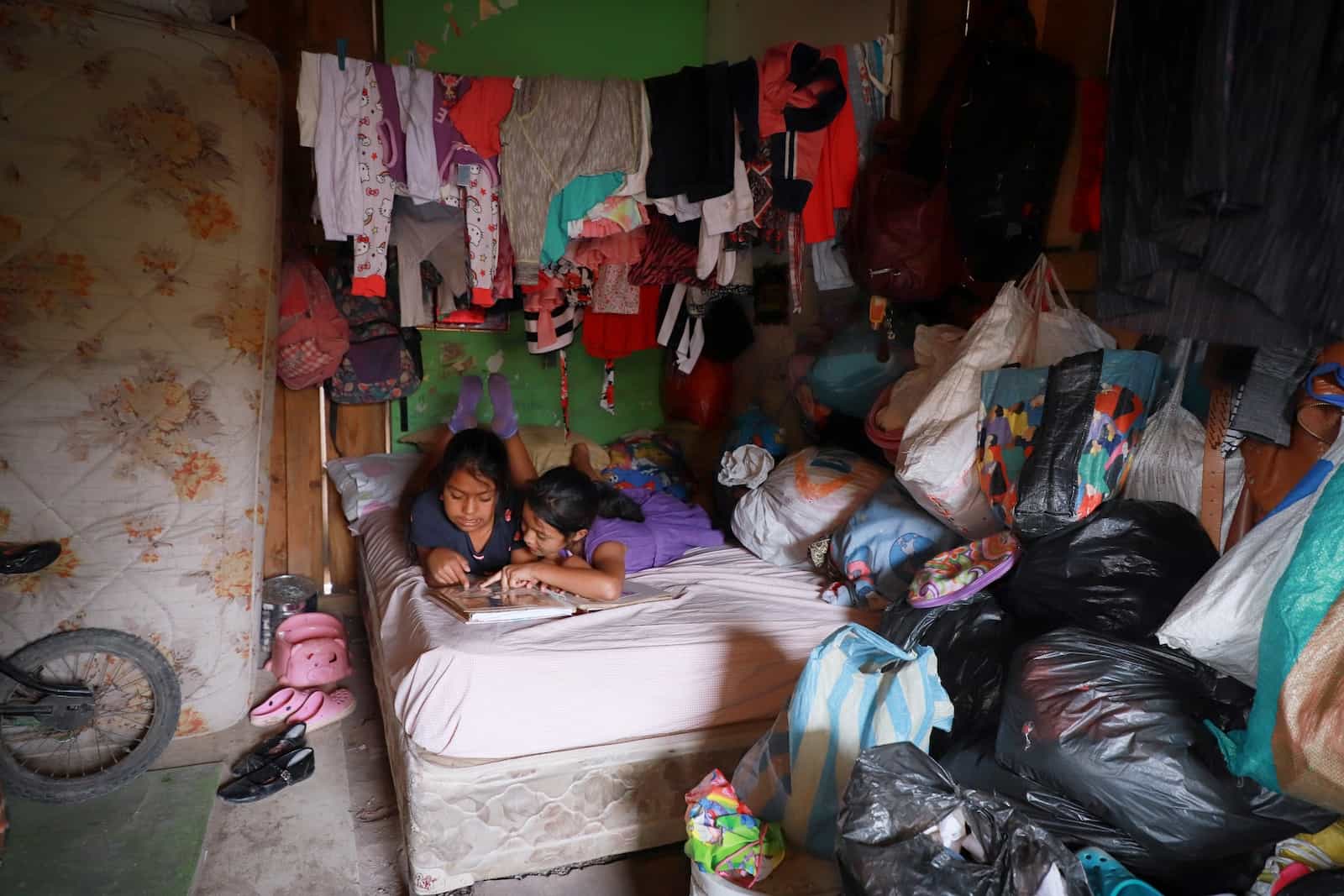 Two girls lie in a bed in a corner of a room, reading a Bible. A drying line of clothing hangs from teh ceiling and the walls and floors are covered in plasticbags, used for storage.