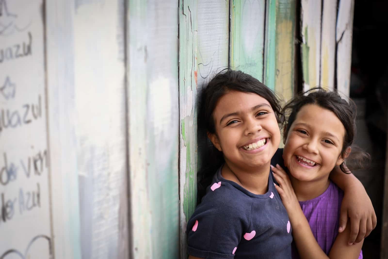 A girl in a blue dress and a girl in a purple dress smile largely at the camera, with their arms around each other, in front of a green wooden wall. 