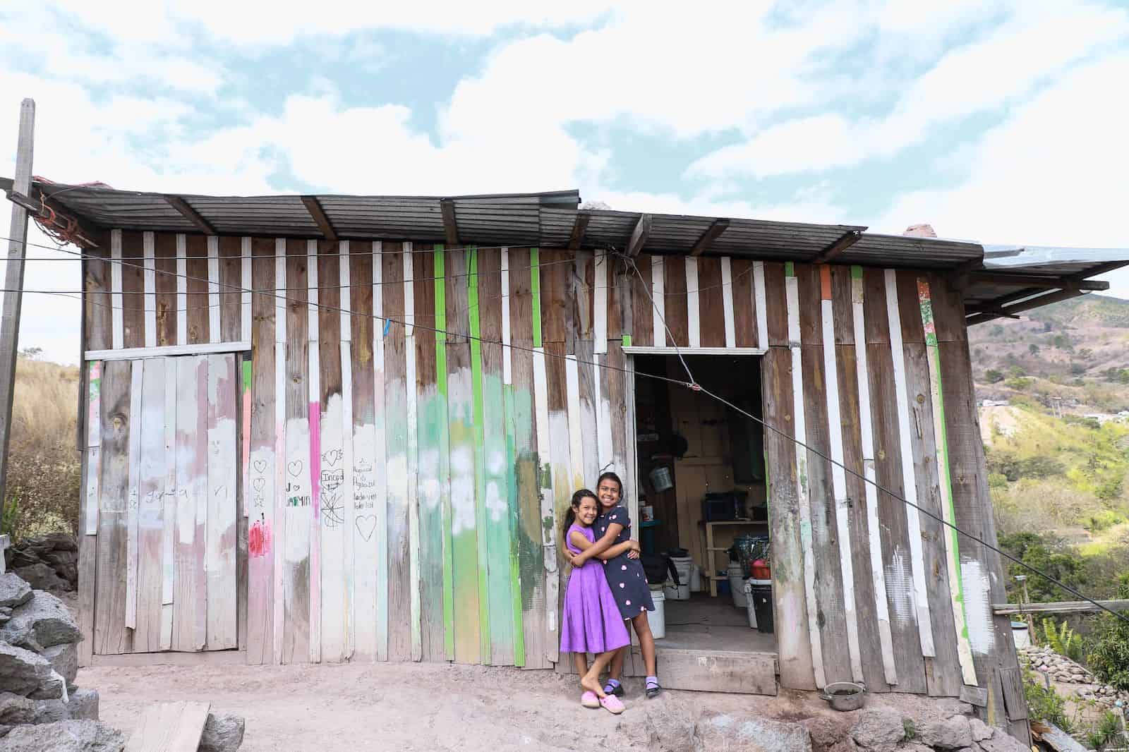 Two smiling girls wearing purple and blue dresses hug outside of a home made from wood and metal sheets, on a hill.