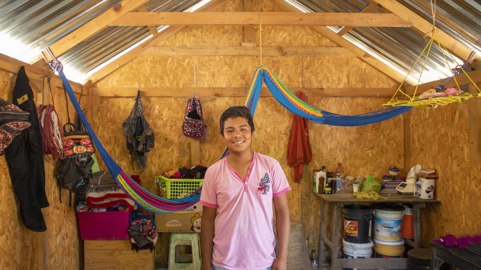 A boy in a pink shirt and jean short stands in the interior of a home built from plywood and metal sheets. Colorful hammocks hang from the ceiling, and personal items, like backpacks hang on the walls. There is a small table in one corner.