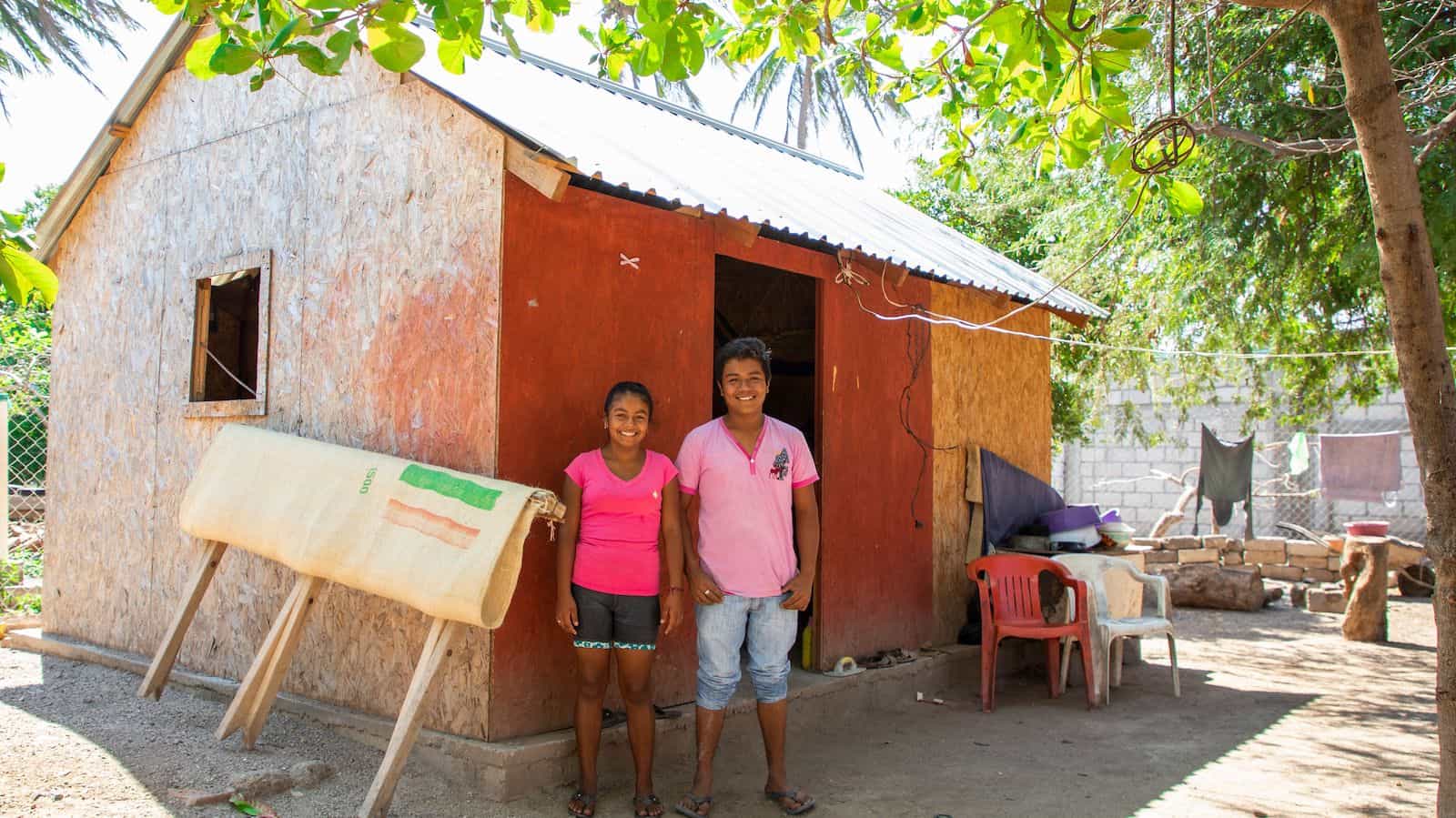 A teenage boy in a pink shirt and jean shorts and a teenage girl in a pink shirt and jean short stand in front of a simple home made of plywood and and metal sheet roof. In the background are trees. 