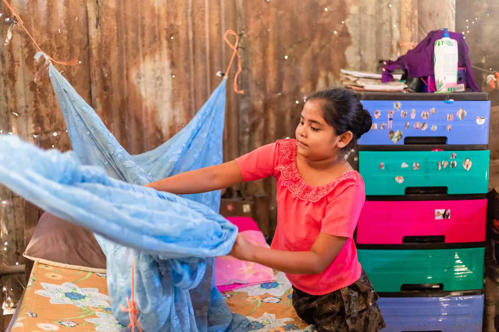 A girl in a pink shirt adjusts a mosquito net above a bed. There is a brightly colored plastic dresser in the background, in front of worn corrugated metal walls. 
