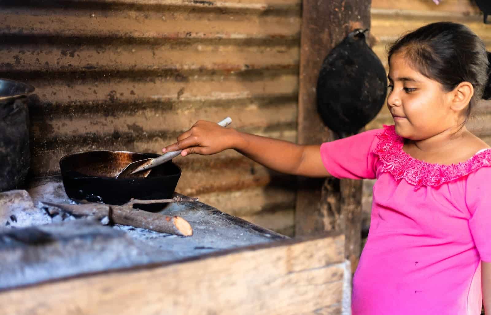 A girl in a pink shirt stands in front of a wooden block with a black pan on it, stirring something in the pan with a spatula. the wall is made of corrugated metal sheets.