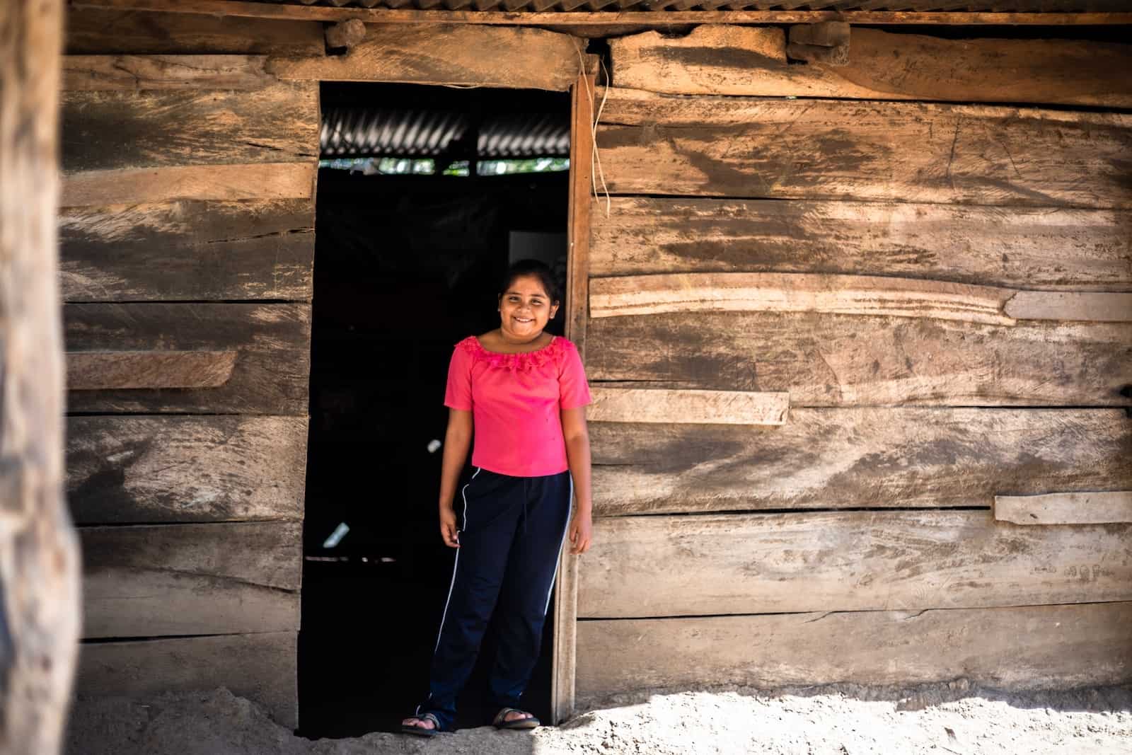 A girl in a pink shirt and jeans stands in the doorway of a wooden home in Central America. 
