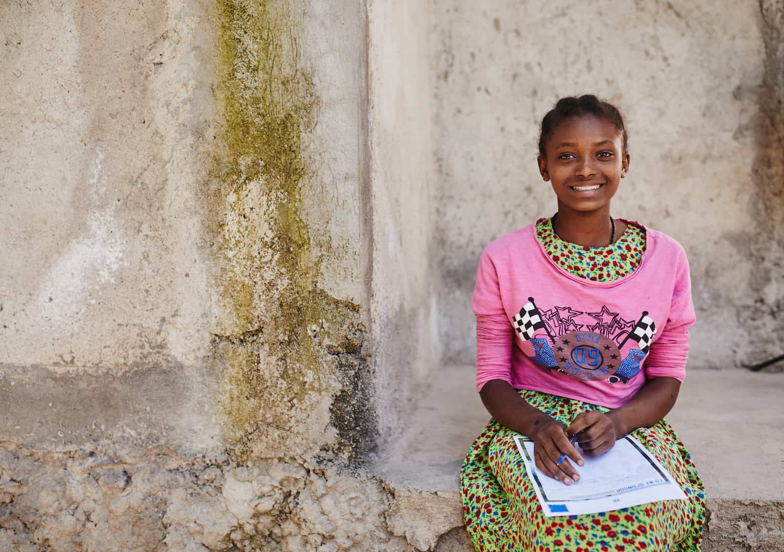 A girl in a pink shirt over a green patterned dress smiles at the camera, sitting on a concrete sidewalk. In her lap, she holds a pen and letter. 