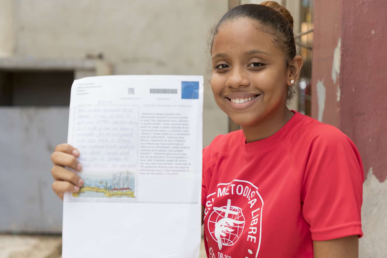 A girl in a red T-shirt smiles and holds up a letter. She sits in front of a concrete wall and wooden post. 
