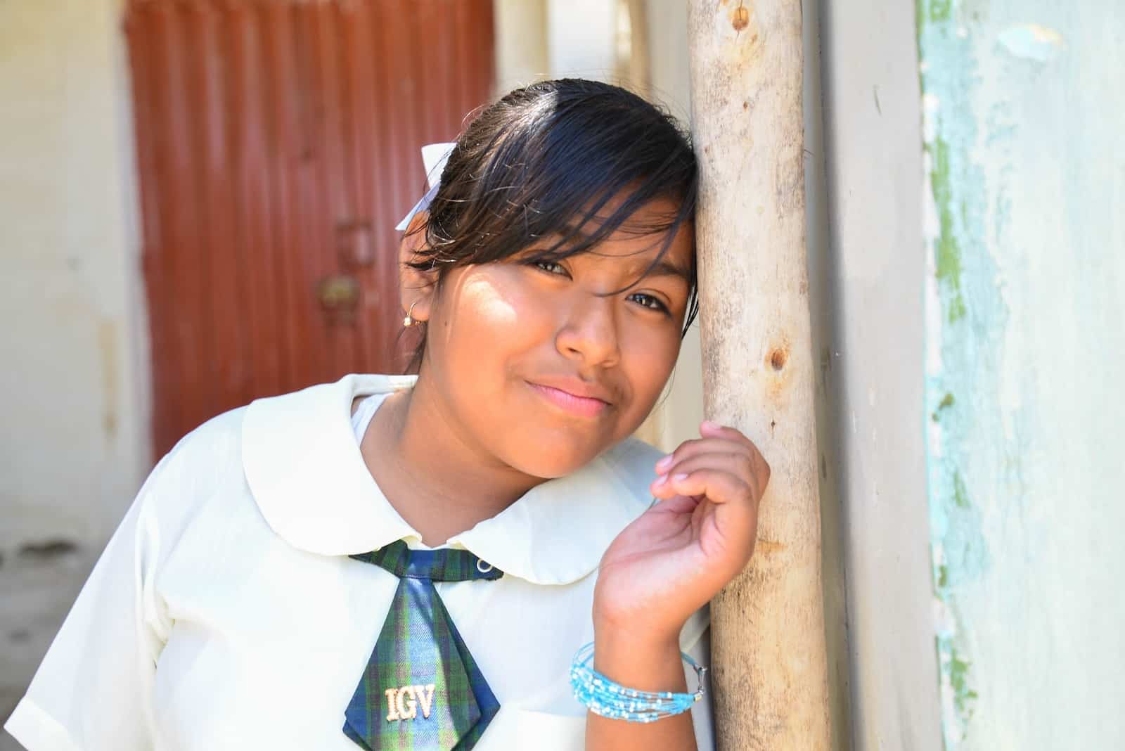 A girl wearing a white button-up shirt and green tie leans her head against a concrete wall outside and smiles at the camera. 