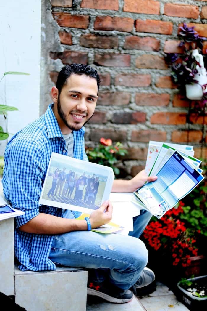 A young man wearing a blue shirt and jeans sits, holding letters and photos, smiling at the camera. In the background is a brick wall and flowers in a pot.