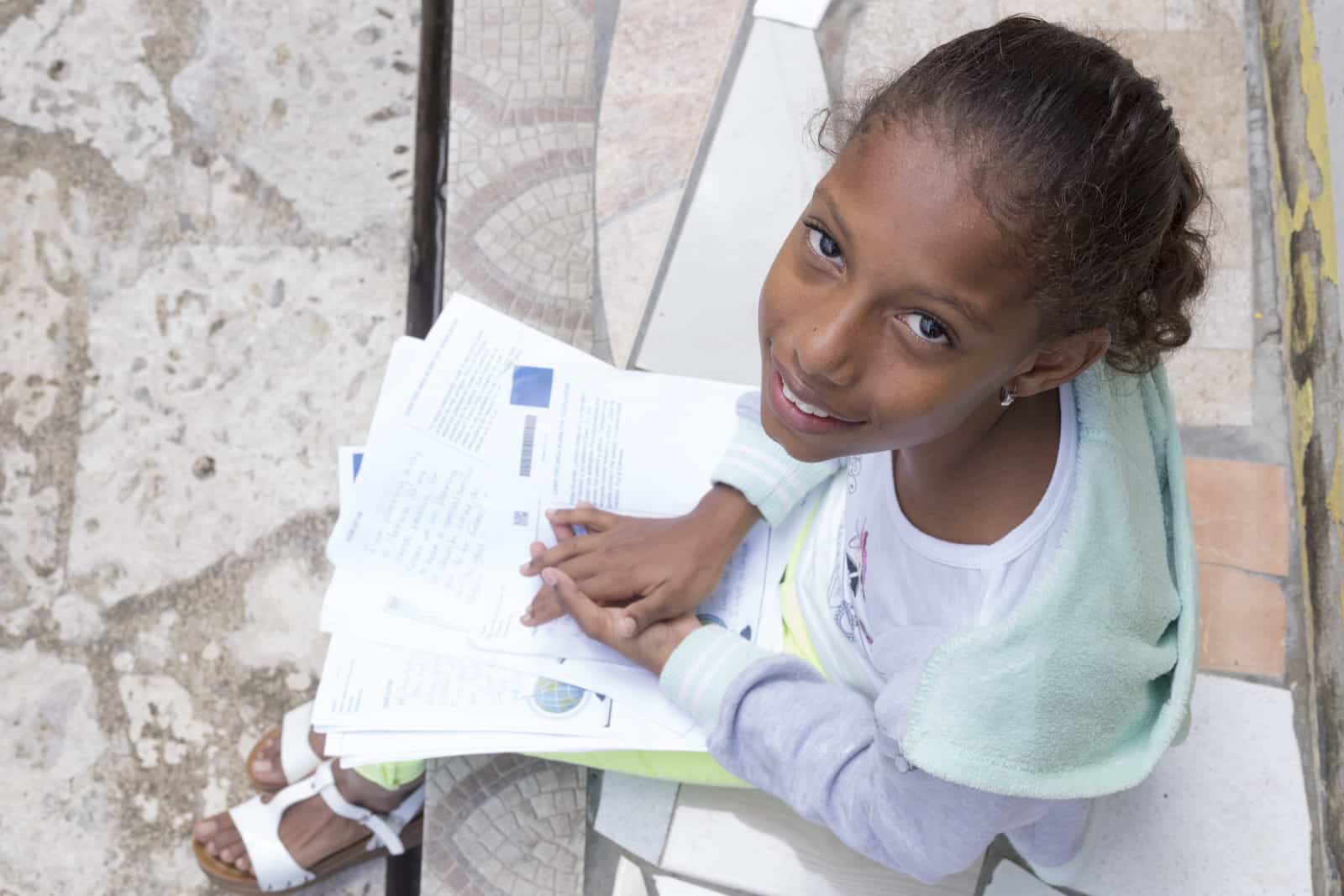 A girl wearing a grey and green sweatshirt sits with her hands folded on top of a pile of letters, as she looks up at the camera.