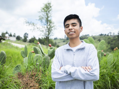 A teenage boy wearing a grey sweater stands with his arms crossed, smiling, in front of a green field.
