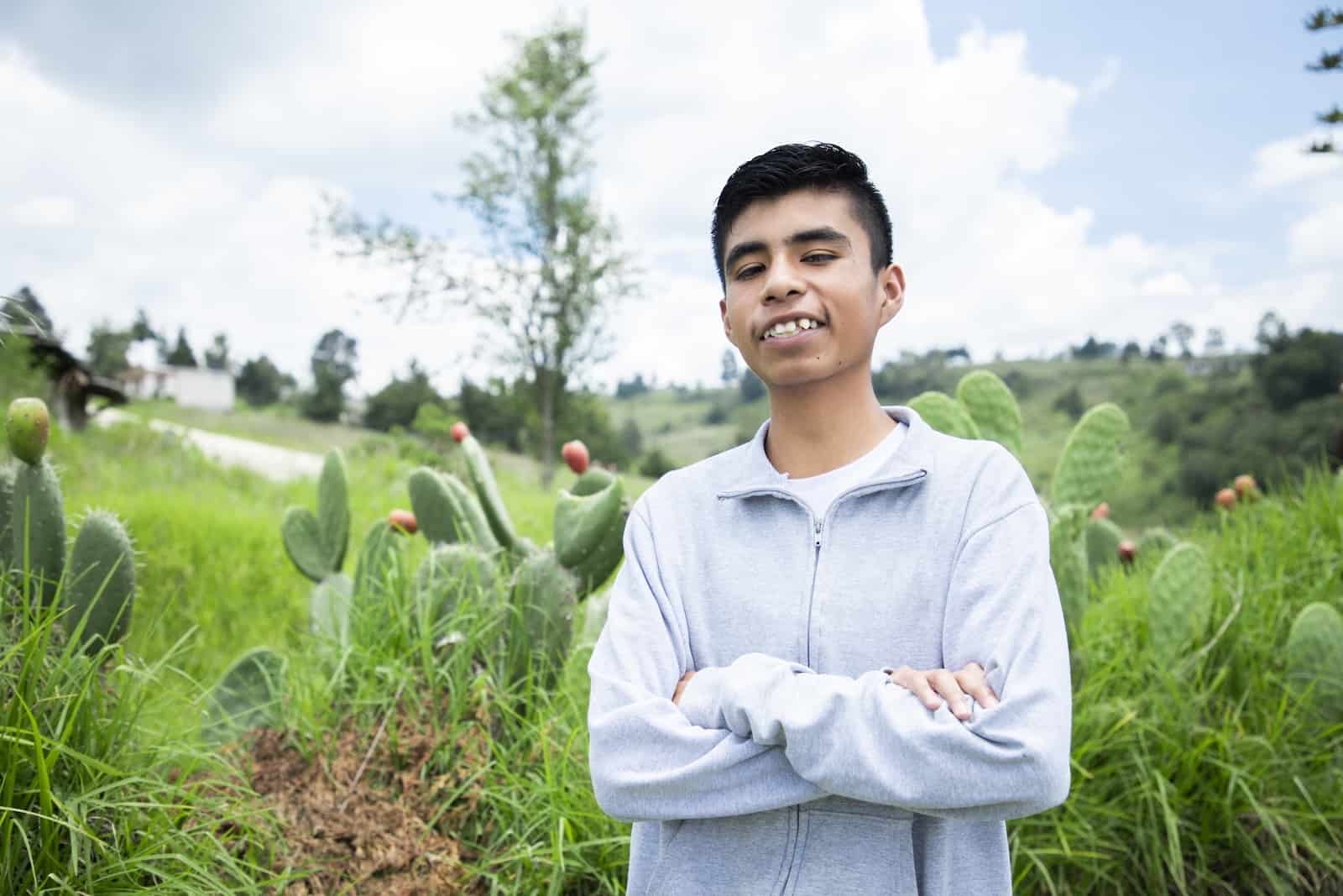 A teenage boy wearing a grey sweater stands with his arms crossed, smiling, in front of a green field.