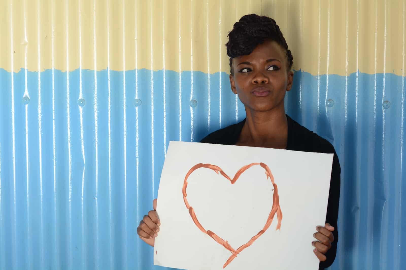 A young woman leans against a blue and white corrugated metal wall holding a white sign with a heart drawn on it. 