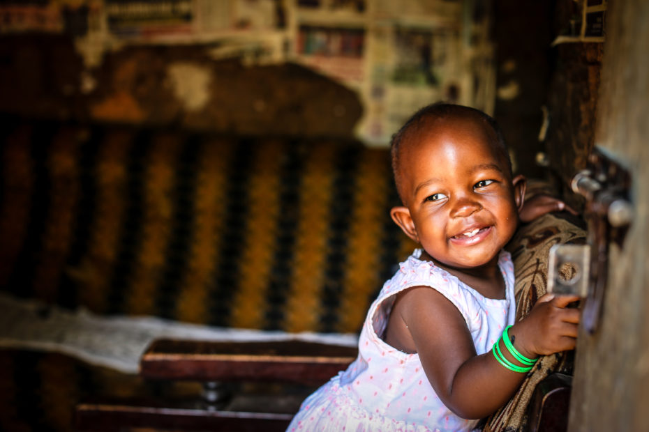 A baby in a white dress leans on the back of a chair and smiles, looking to the side. In the background is a brown couch and a wall covered in newspapers.