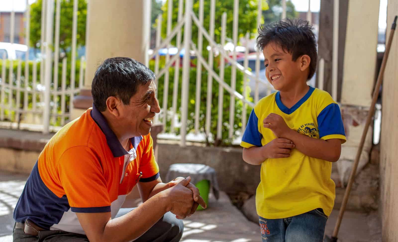 A man in an orange shirt kneels next to a boy in a yellow shirt, talking to him in. 