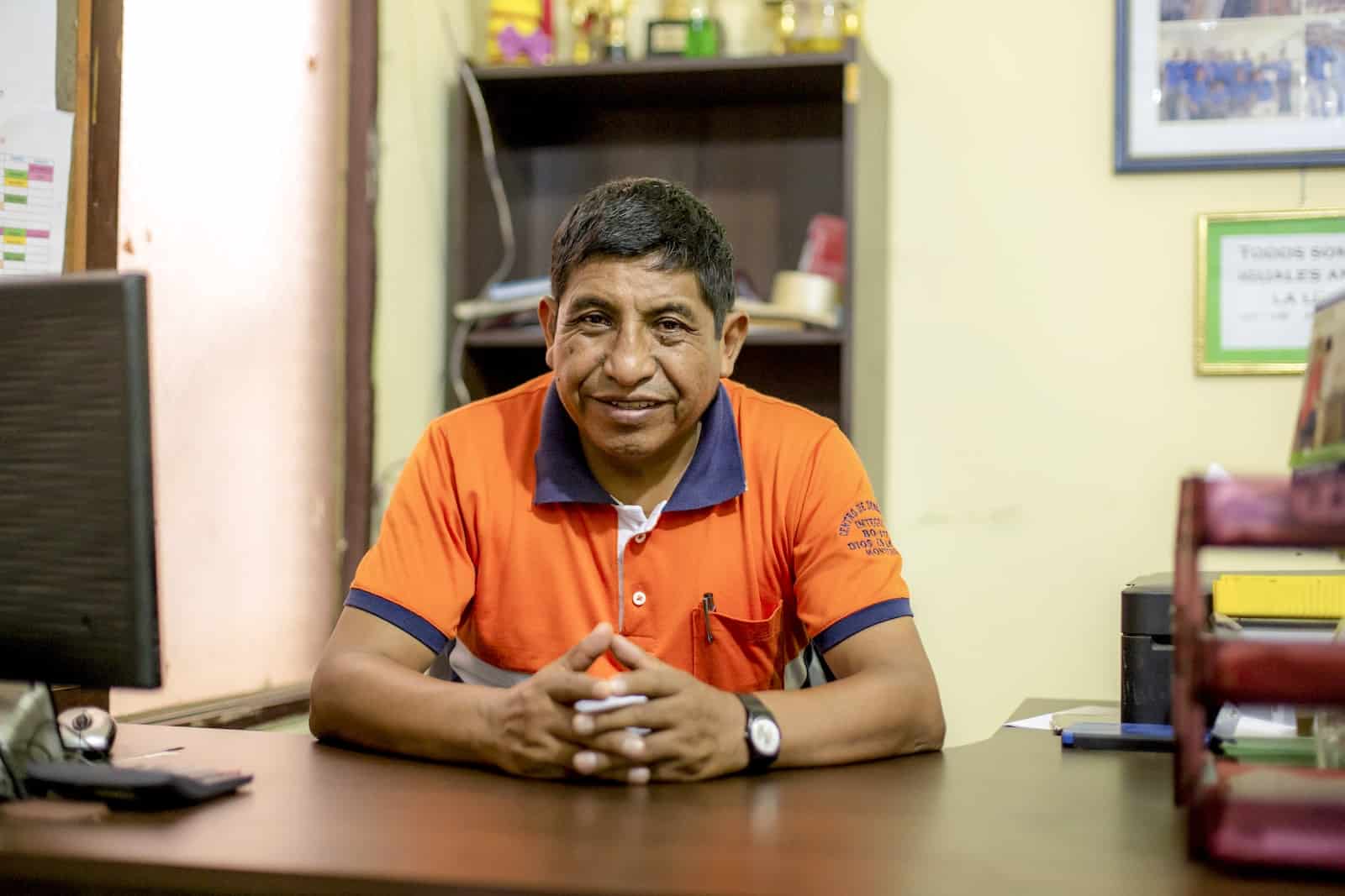 A man in a short sleeved orange shirt clasps hands in front of him, sitting at a desk. 