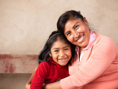 A woman in a coral colored sweater hugs a little girl in a red shirt.