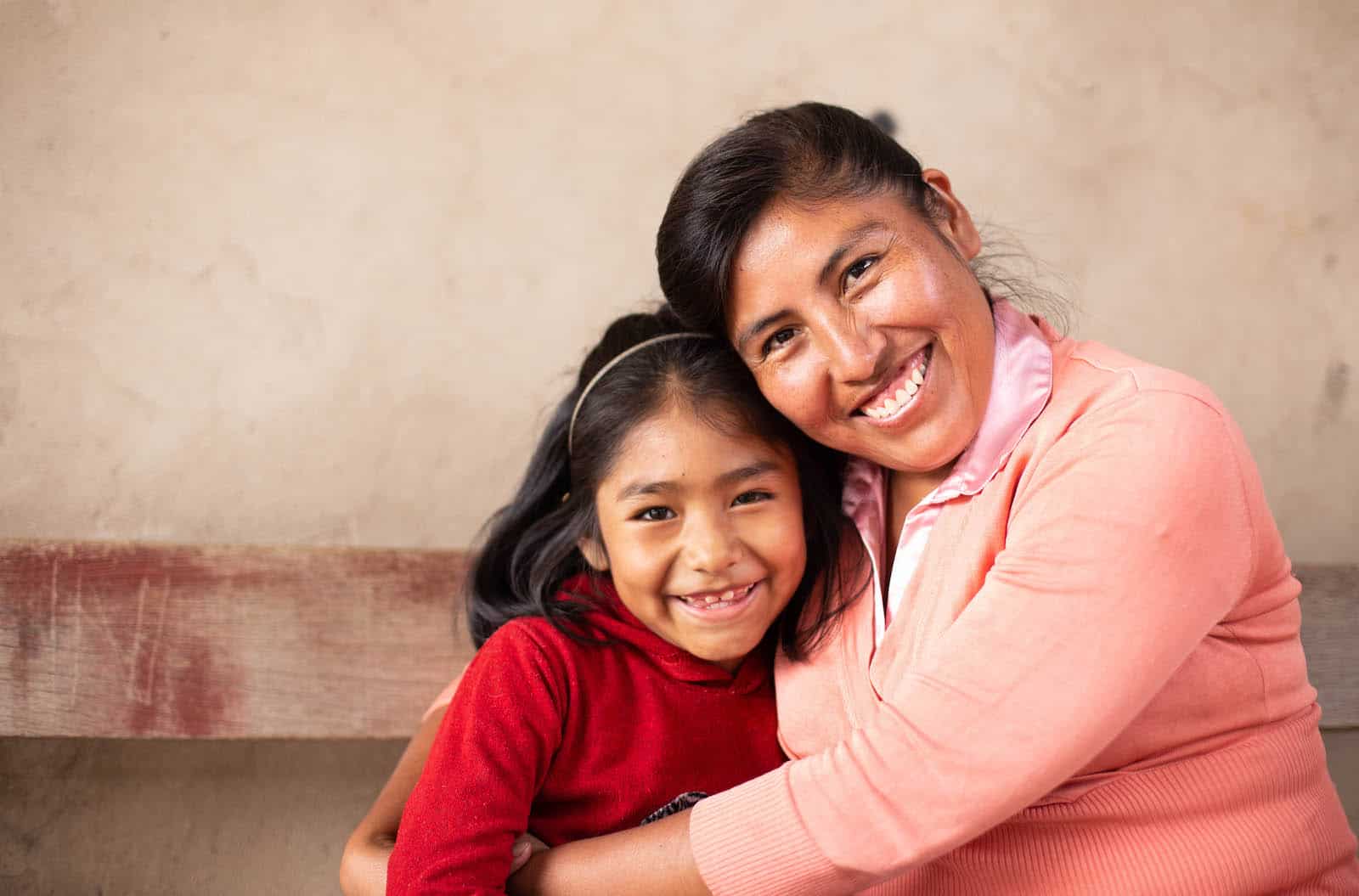 A woman in a coral colored sweater hugs a little girl in a red shirt. 