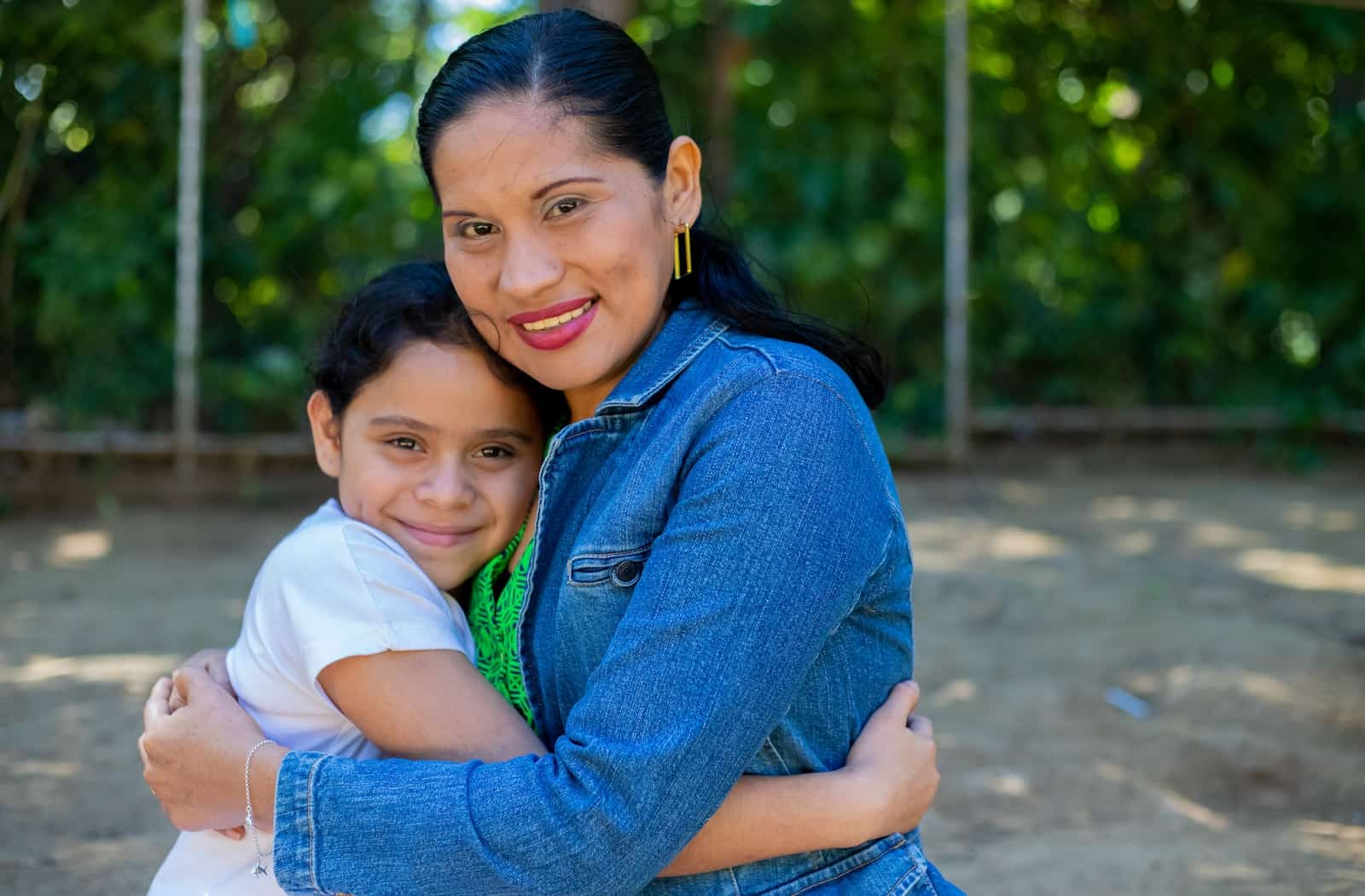 a woman in a blue shirt hugs a young girl in a white shirt outside. 