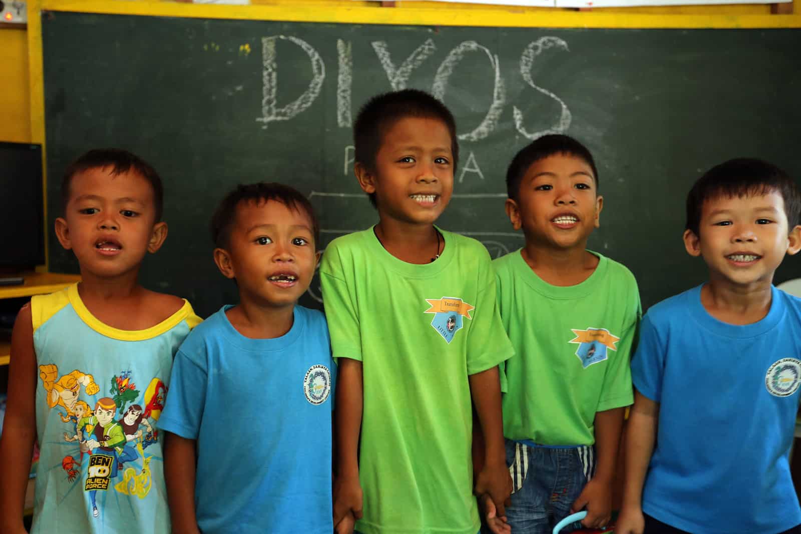 A group of five young boys stand, smiling, in front of a chalk board.