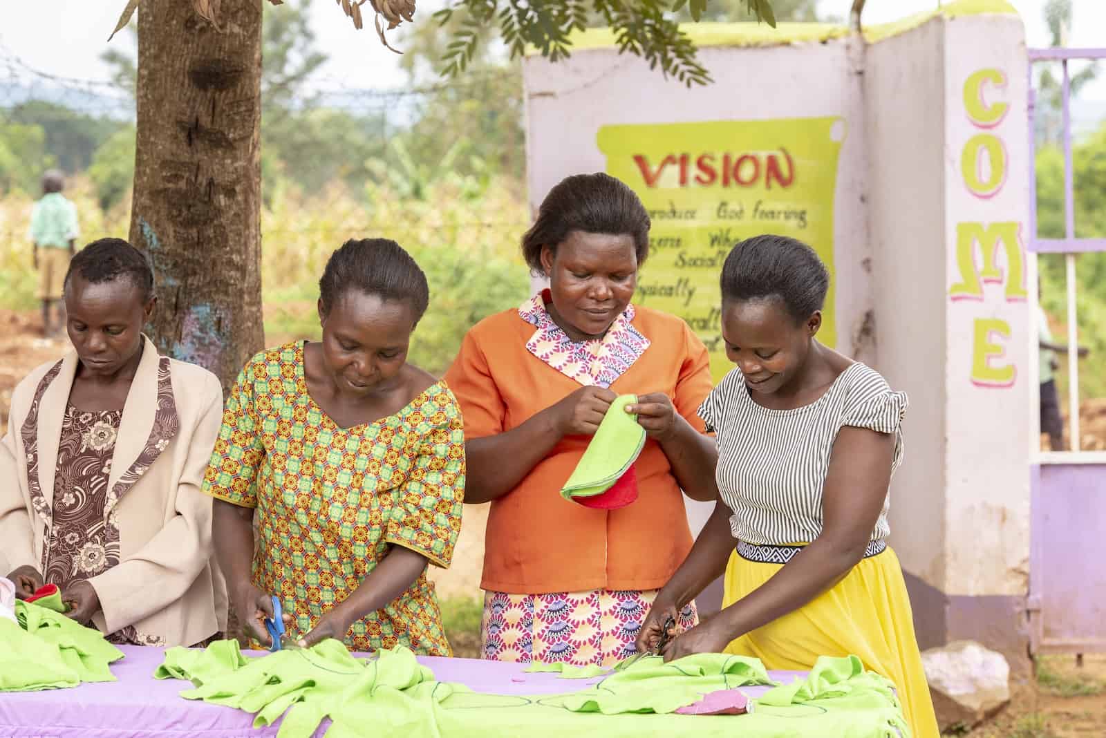 A group of four women stand behind a table, working with green pieces of fabric. They are outside with a palm tree in the background. 