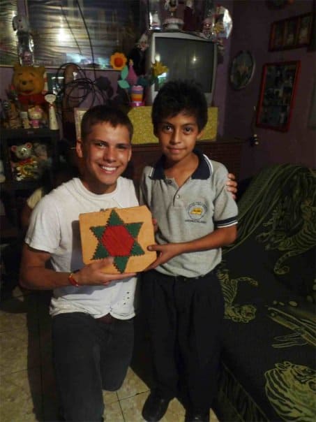 A teenage boy kneels net to a boy as they hold a pillow with a star on it together. They are inside a dark home. 