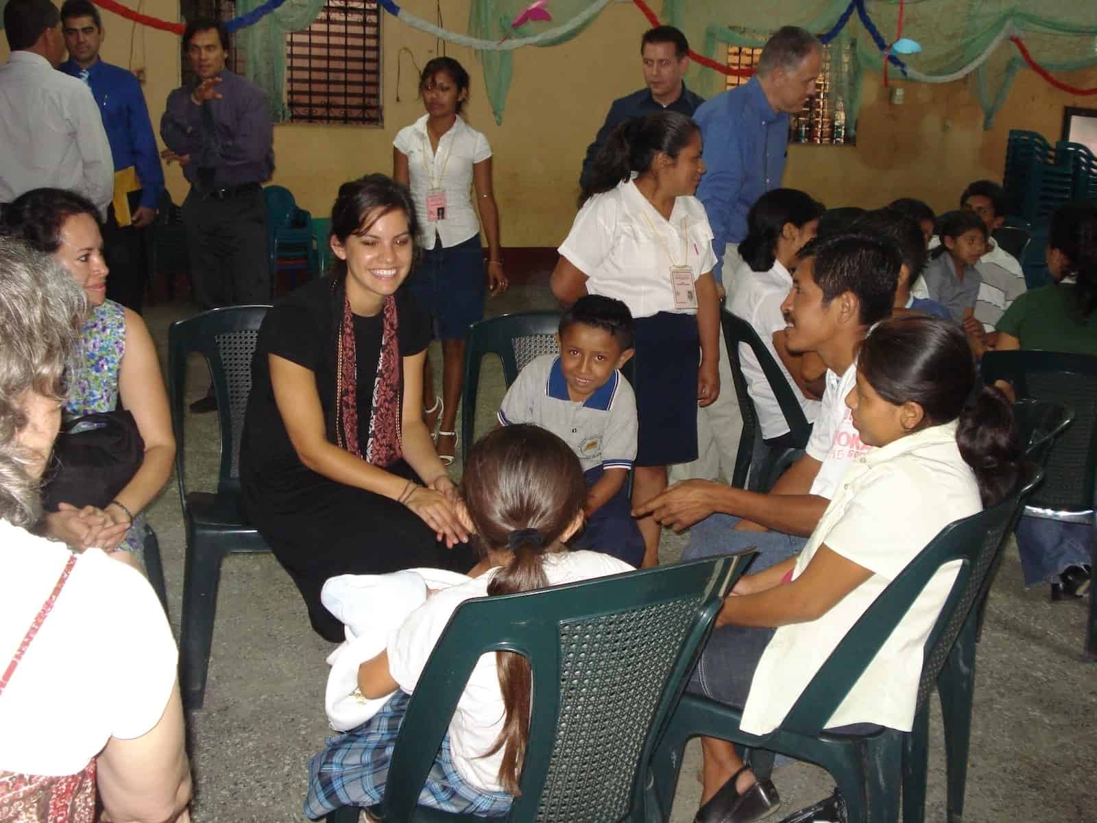 A girl sits in a circle of chairs, talking to children inside a church room.