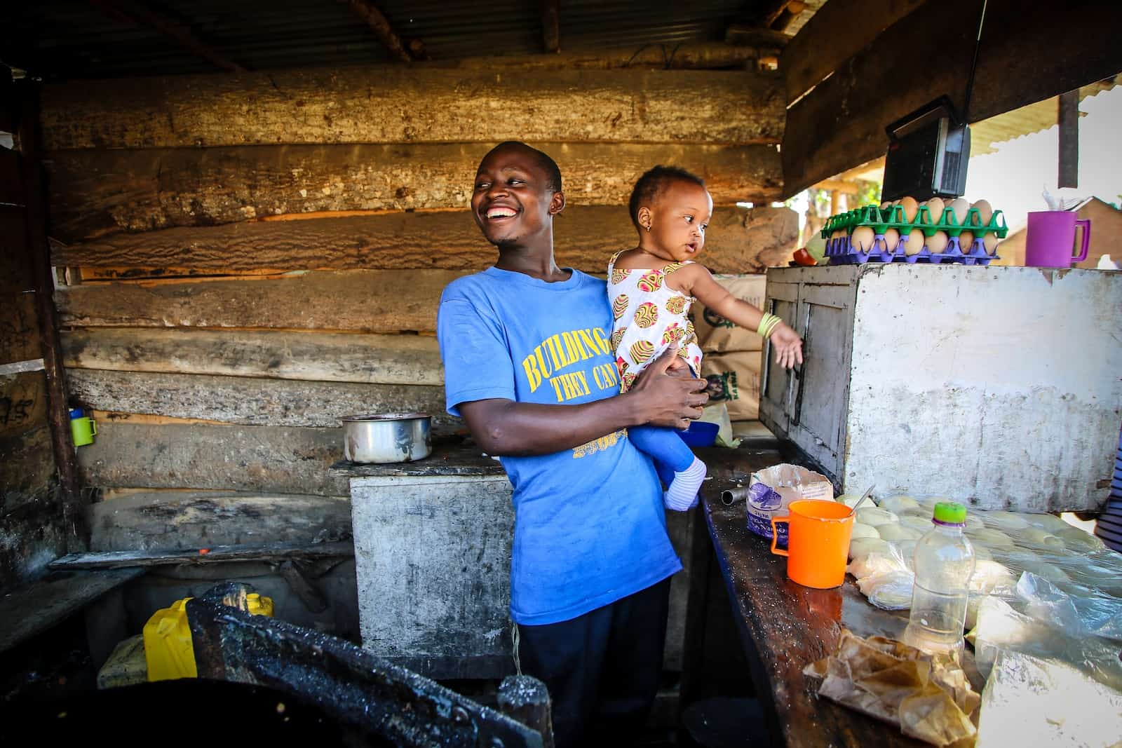 A man wearing a blue T-shirt holds a baby girl in his arms, standing in an open-air kitchen with breads and eggs on a table.