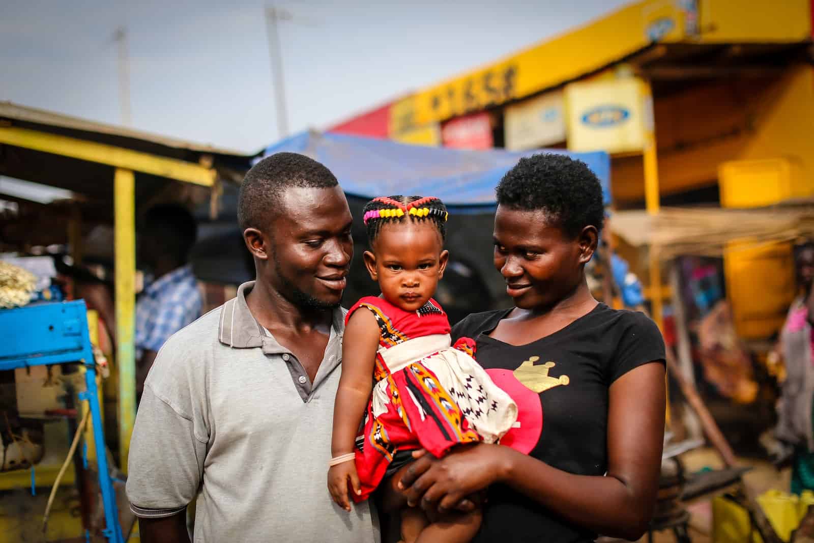 A man in a grey shirt stands next to a woman in a black shirt holding a small girl in a red and white dress. They are standing on a street, both looking at the girl. 