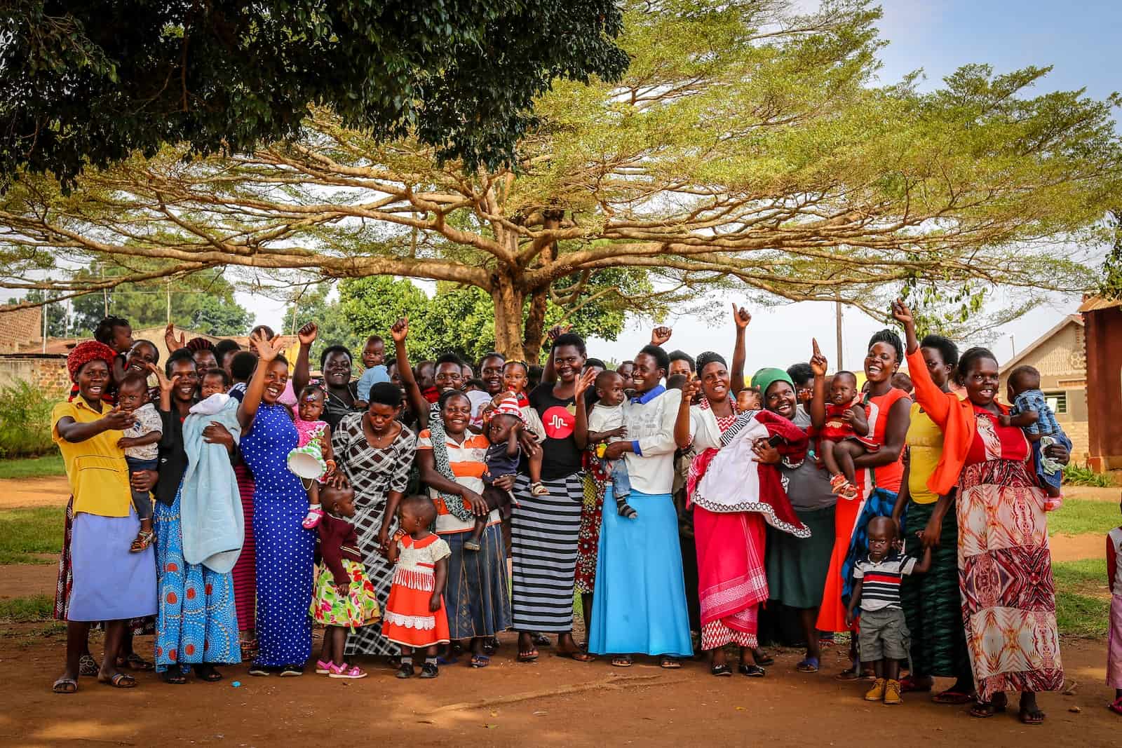 A group of women wearing long dresses and holding babies stands under a tree and cheer, some raising their hands in the air, celebrating an important birthday milestone.