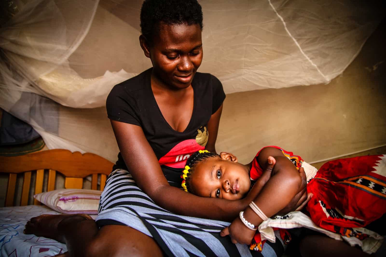 A girl wearing a red and white dress lays on her mother's lap. The mom wears a black shirt and black and white striped dress. They sit on a bed with a mosquito net on it.